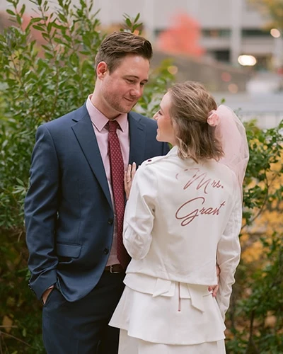 A bride and groom smile at each other outdoors. The back of the bride's white jacket reads 