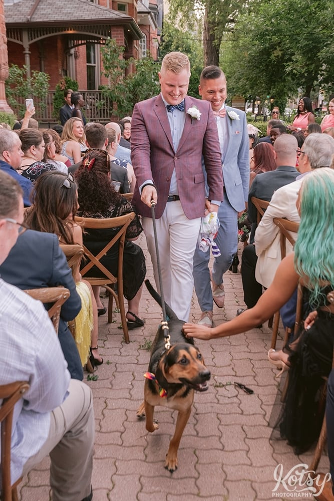 Two grooms walk up the aisle with their dog after their outdoor wedding ceremony at Berkeley Bicycle Club in Toronto, Ontario