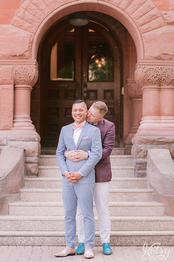A groom gives his groom a kiss from behind in front of Berkeley Bicycle Club in Toronto