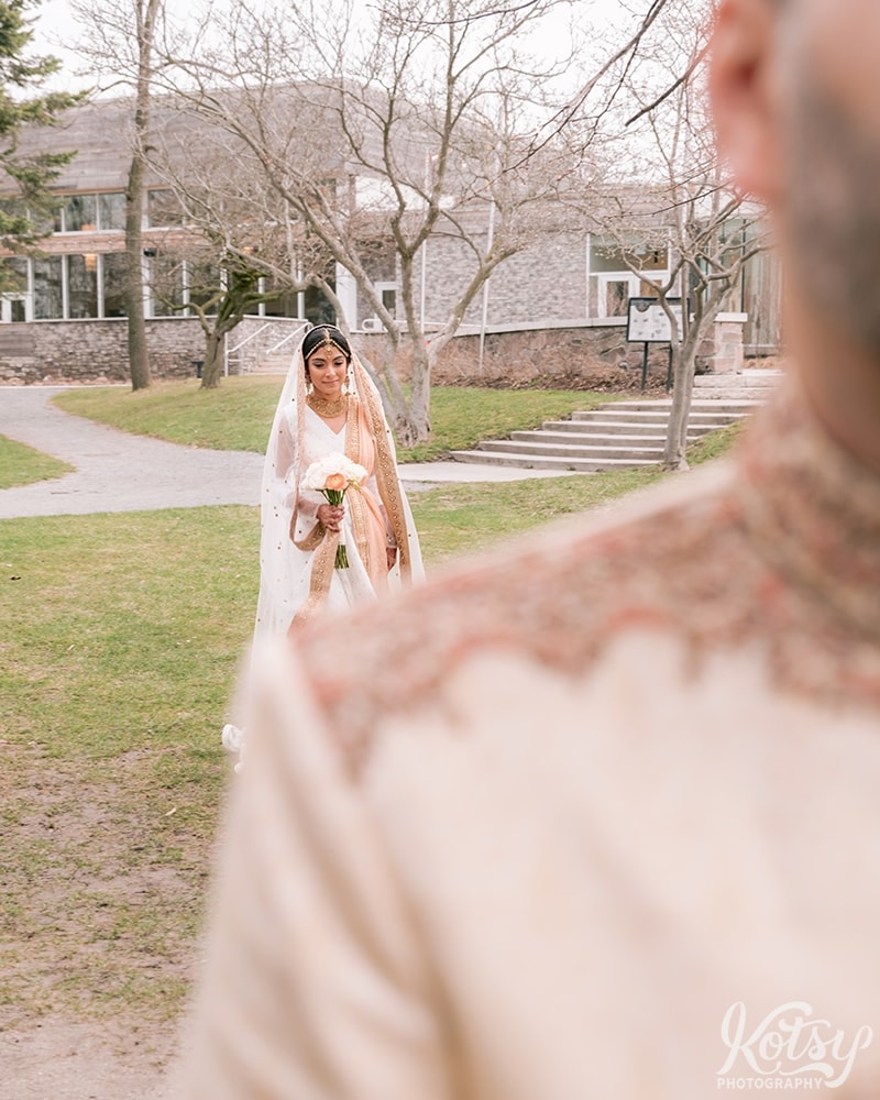 A bride is seen over a groom's shoulder as she walks to a first look photo session. Photographed at Guild Park and Gardens in Scarborough.