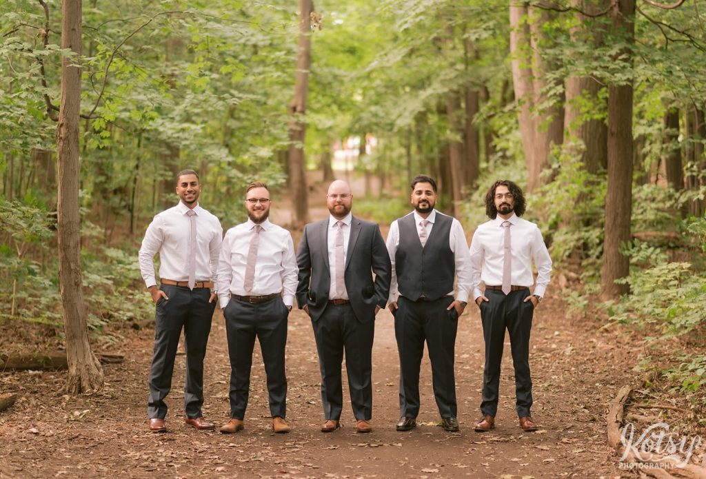 A groom and his groomsmen pose for a group photo in the on a dirt path through a forest.