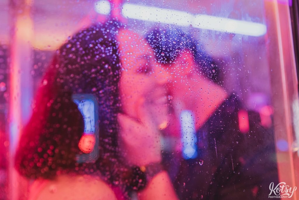 A man kisses his girlfriend's cheek behind a rain-drop glass partition of a phone booth at Neon Demon Studio in Toronto