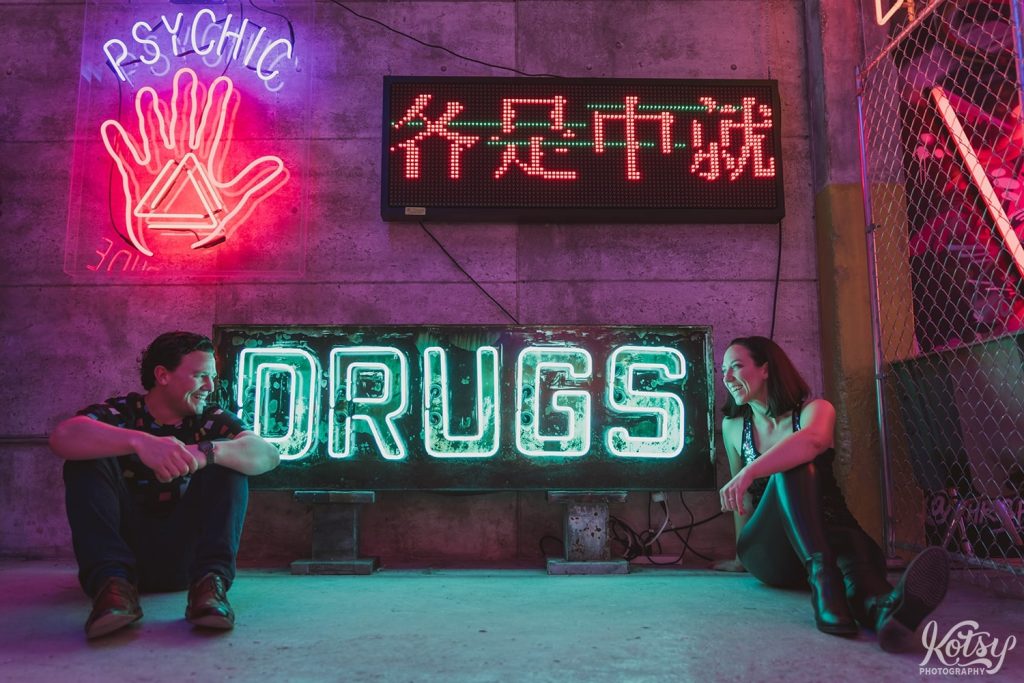 A recently engaged couple sit next to a neon 'drugs' sign at Neon Demon Studio in Toronto