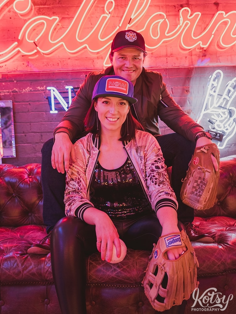 A recently engaged couple sit on a leather couch in ball caps and baseball gloves on at Neon Demon Studio in Toronto, Ontario