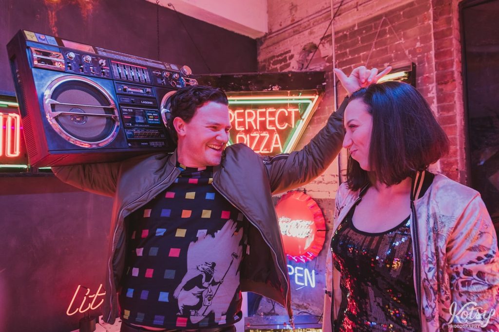 A excited man smiles ear to ear while holding a ghetto blaster while his new fiancé pouts. Photographed at Neon Demon Studio