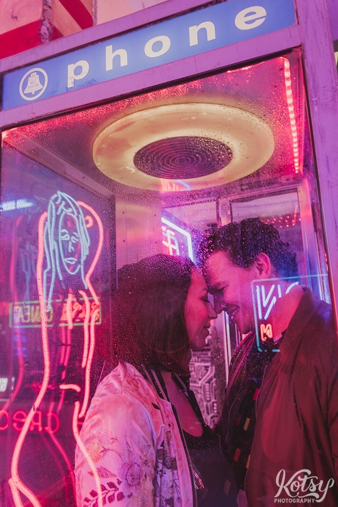 A recently engaged couple stand forehead to forehead inside a phone booth with rain drops on the glass. Photographed at Neon Demon Studio in Toronto, Ontario