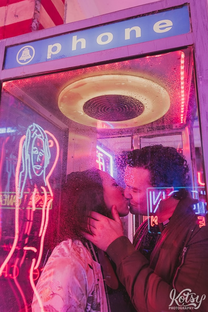 A recently engaged couple kiss inside a phone booth with rain drops on the glass. Photographed at Neon Demon Studio in Toronto, Ontario