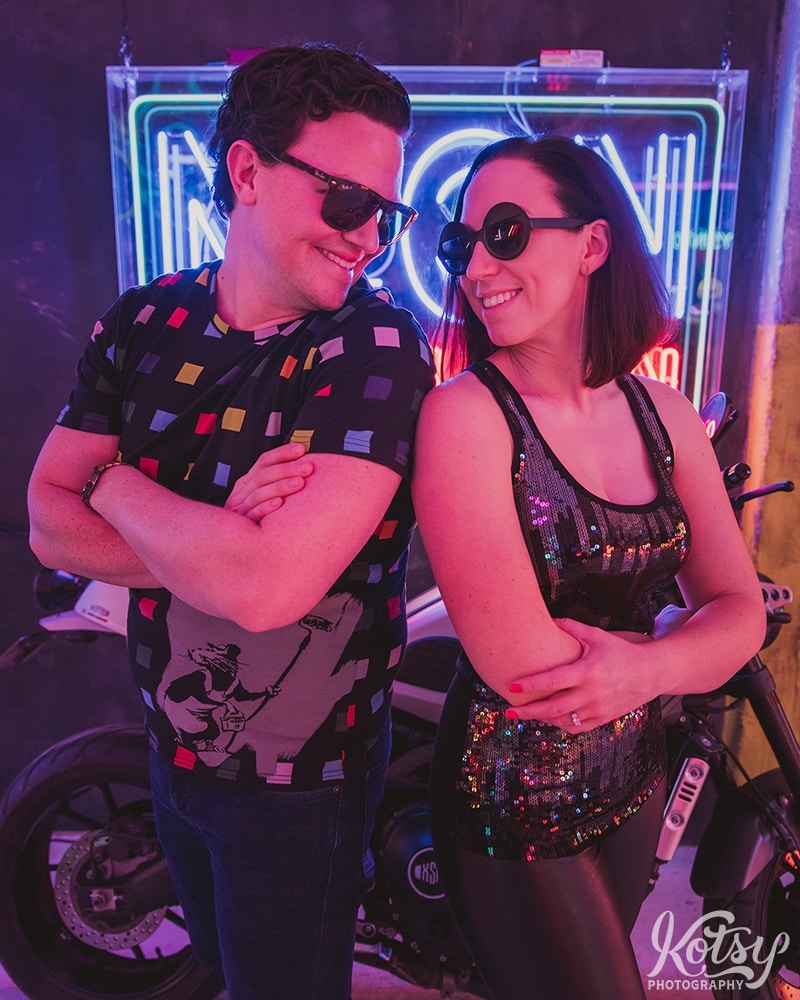 A recently engaged couple stand at an angle shoulder-t-shoulder with dark sunglasses on while grinning at each other. Photographed at Neon Demon Studio in Toronto