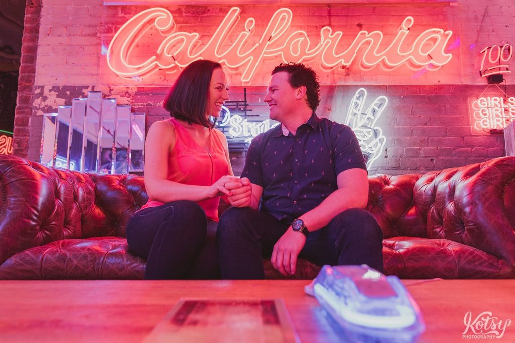 A recently engaged couple sit on a leather couch smiling at each other. A neon California sign is seen on the brick wall behind them with a neon lit phone in the foreground.