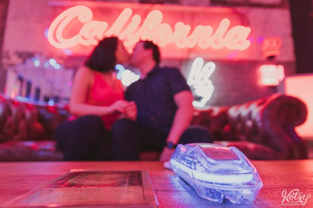 A recently engaged couple embrace each other on a leather couch smiling at each other. A neon California sign is seen on the brick wall behind them with a neon lit phone in the foreground.