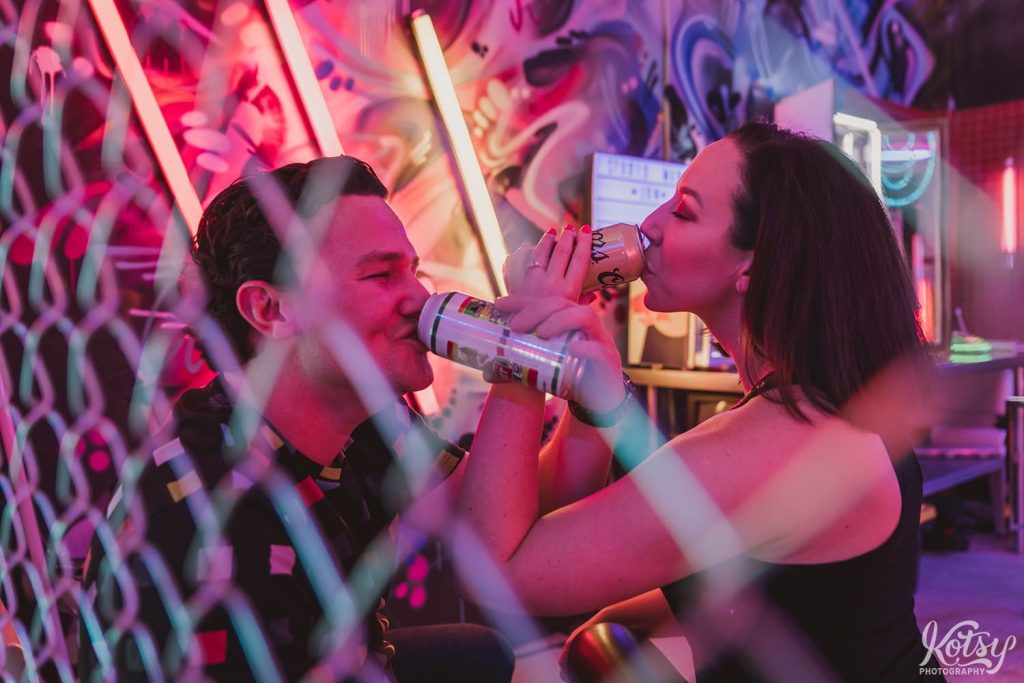 A recently engaged couple chug a beer with arms interlocked behind a chain link fence at Neon Demon Studio.