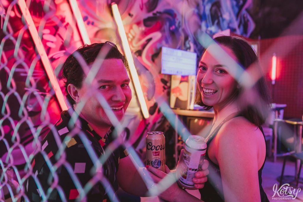 A recently engaged couple smile behind a chain link fence at Neon Demon Studios in Toronto