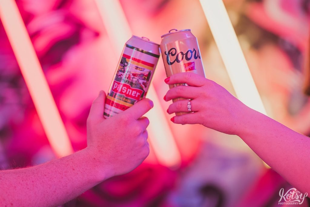 Two beer cans are cheers by a recently engaged couple at Neon Demon Studio