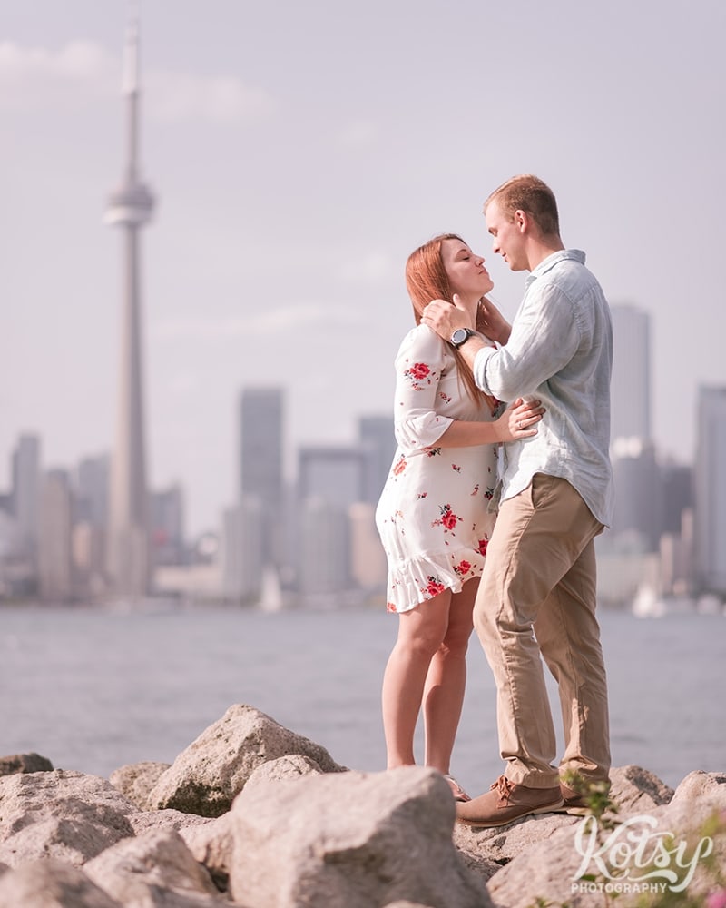 A man goes in for a kiss with his fiancé on some rocks on the Toronto Islands. The downtown skyline is seen in the background.