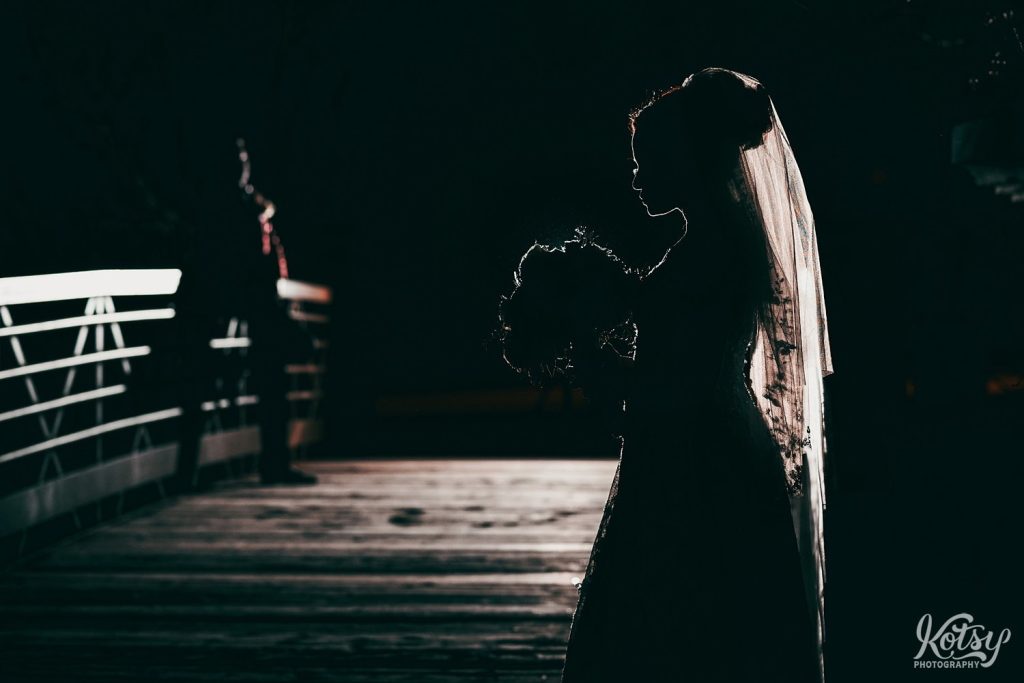 A bride's silhouette is seen at nighttime on a bridge with her new husband in the background. Photographed in Brampton, Ontario, Canada