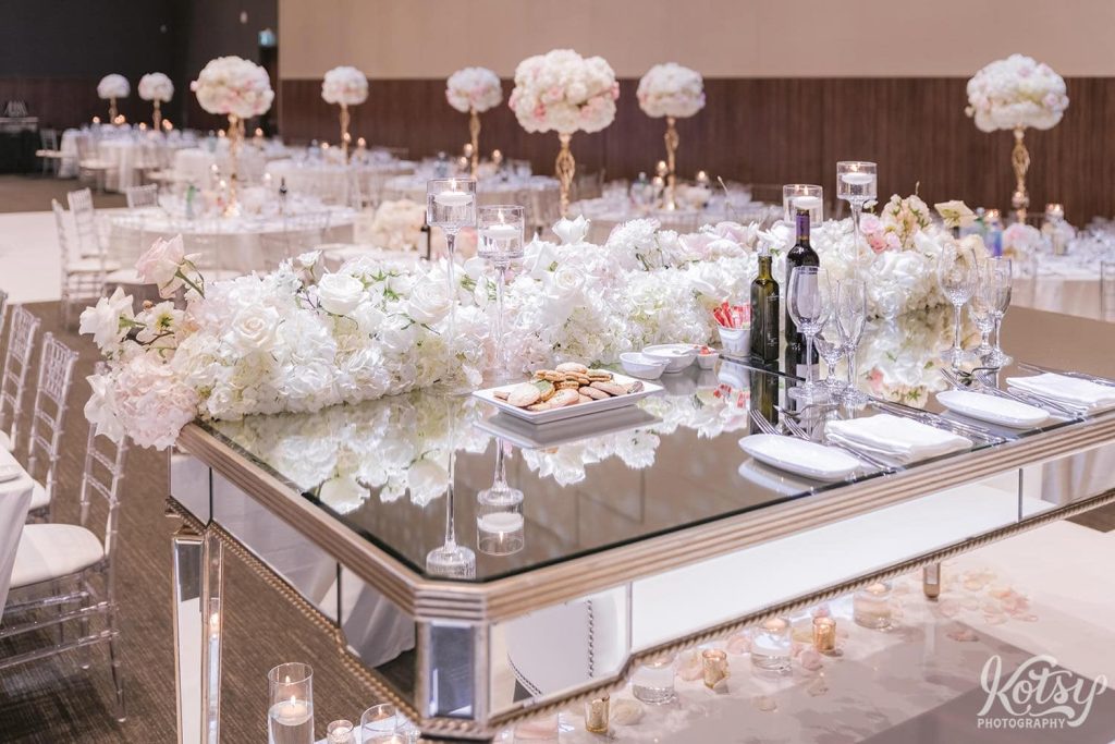 The head table with a mirrored top and flowers are seen in a large wedding venue.