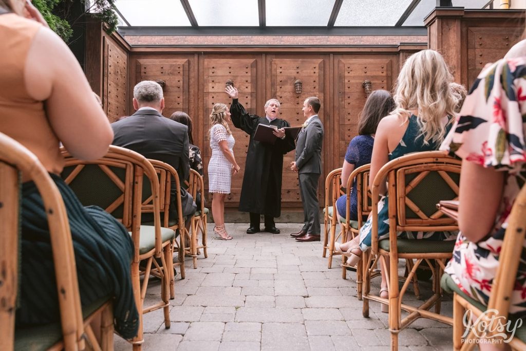 A shot down the aisle with a bride and groom smiling at an officiant waving his hand as he speaks