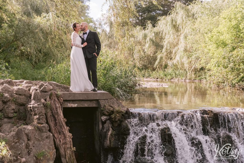 A groom kisses a bride's cheek on a concrete platform next to a waterfall in a park