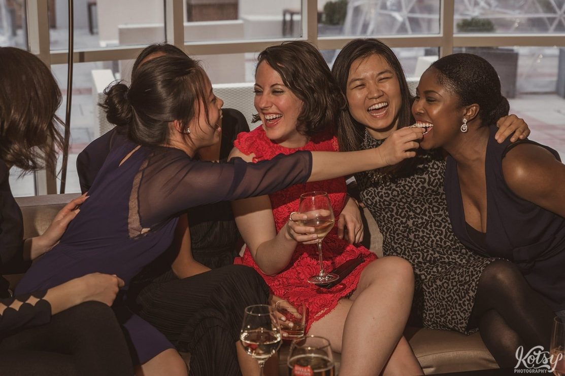 A group of women enjoy big laughs on a couch while sampling food.