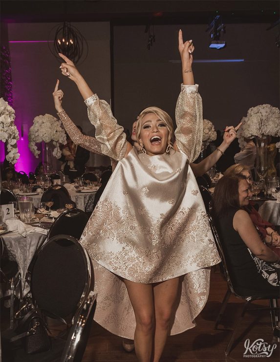 A woman in a white and gold dress struts towards the camera with her hands held high and a big smile on her face.