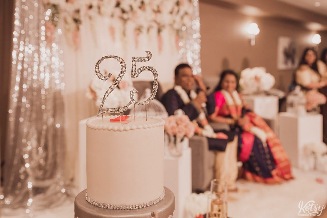 A close up shot of cake topper reading 25. A married couple celebrating their anniversary can be seen out of focus in the background.