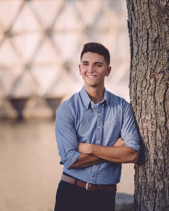 A man poses for a portrait while leaning against a tree in front of the Cinesphere at Ontario Place