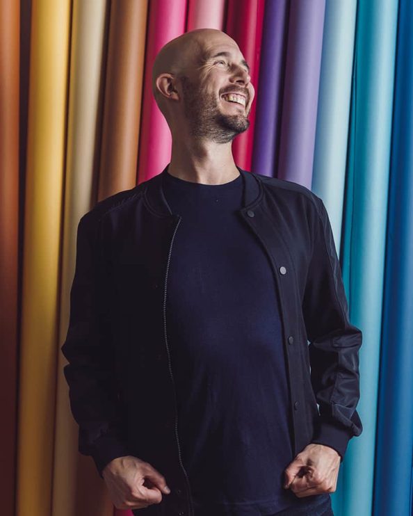 A man lets out a huge smile in front of a collection of coloured paper