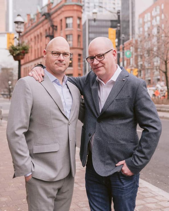 Two men pose for a portrait in front of the Flatiron Building in Toronto