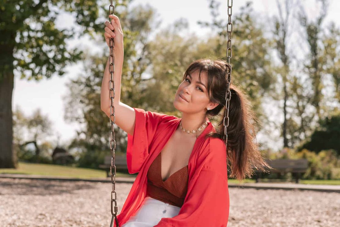 A woman in a red shirt poses for a portrait while sitting on a swing set in Toronto