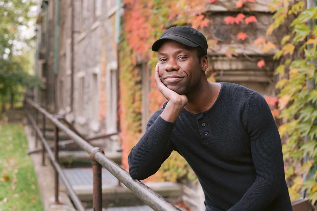 A man poses for a portrait in front of a building covered in fall coloured leaves at University of Toronto