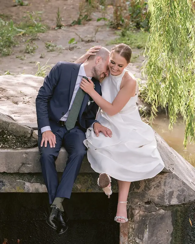 A bride cuddles with her new groom while sitting on a stone platform in a park