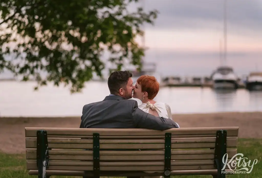 A bride and groom enjoy a kiss at on a waterfront bench at sunset in Toronto, Canada