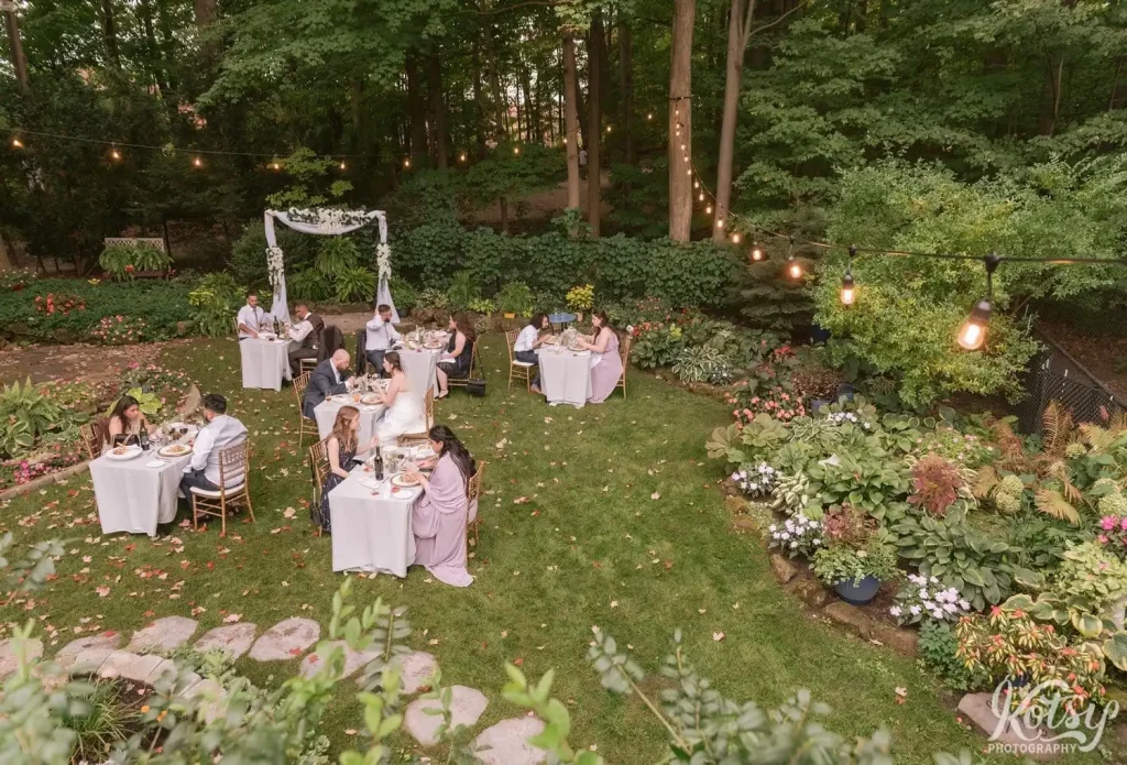 A long string of lights leads down a handful of tables set up in a backyard for a wedding reception