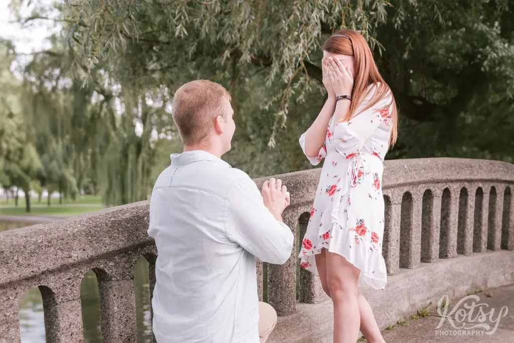A woman covers her face as her partner proposes to her on one knee on the Toronto Islands.