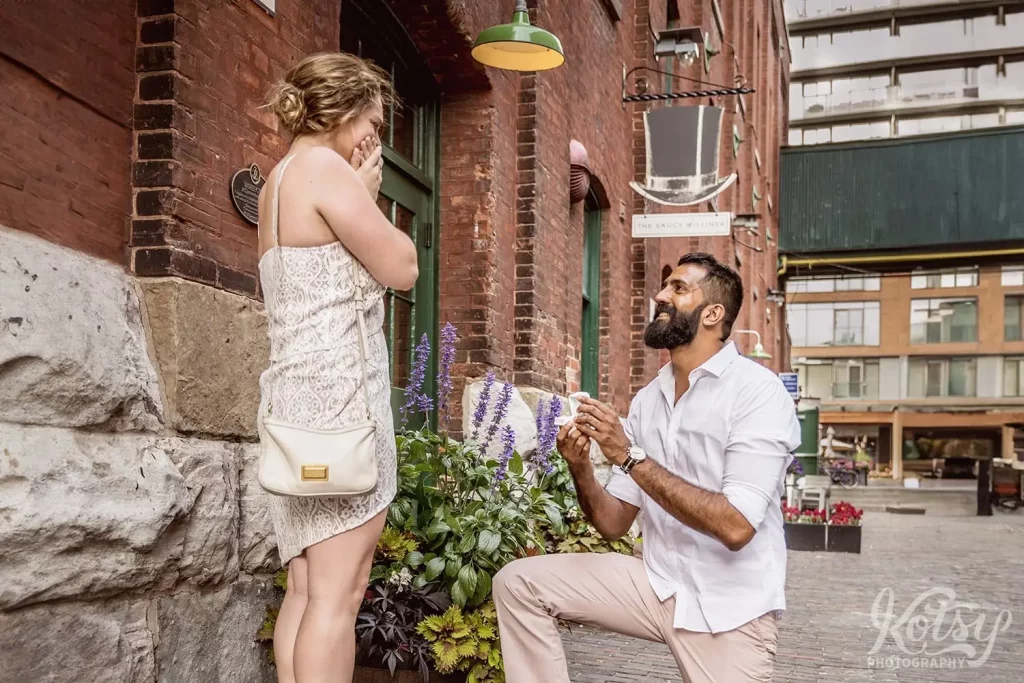 A woman covers her mouth as her parntner proposes to her in Toronto's Distillery District.