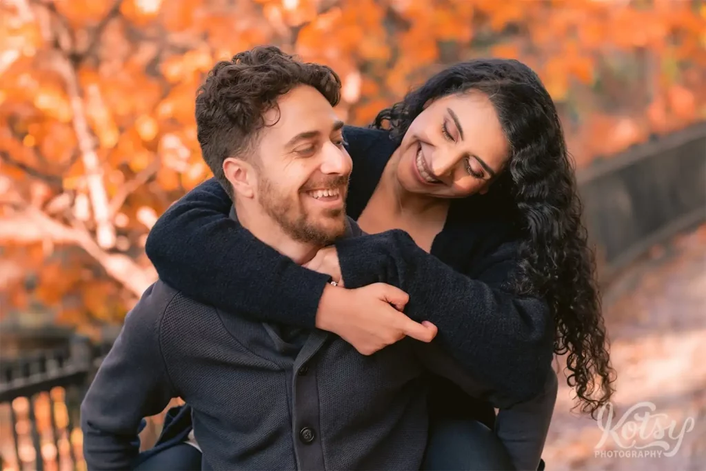 A recently engaged couple enjoy a laugh while piggy back riding through a fall-coloured High Park in Toronto