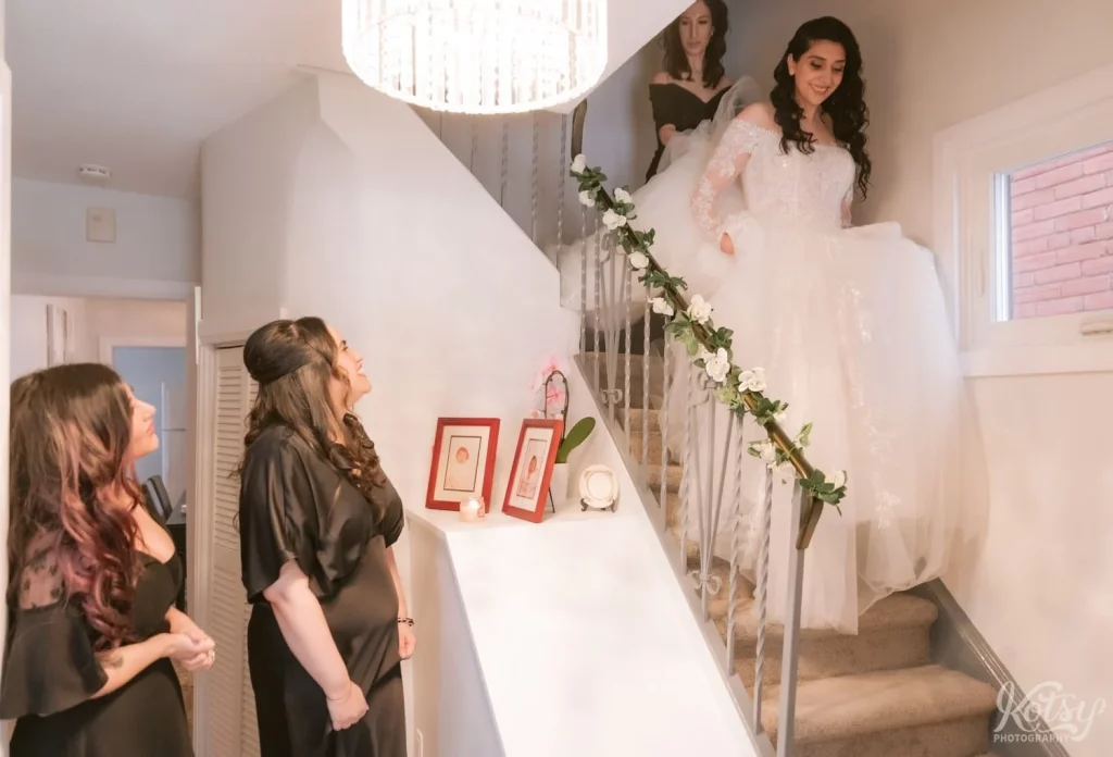 A bride comes down the stairs at her parents house in her wedding gown.