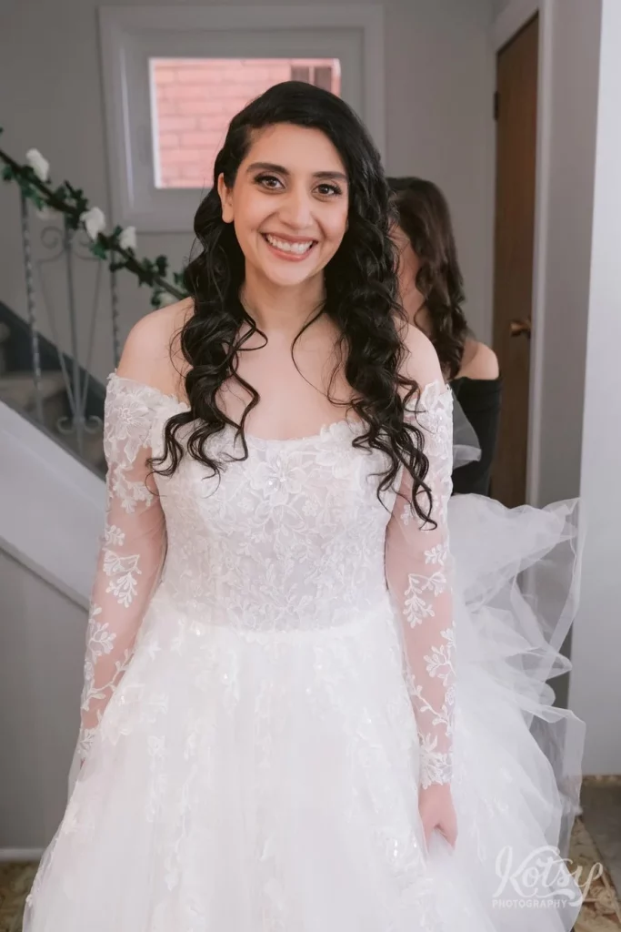 A bride smiles for the camera at her parents house while dressed in her wedding gown.