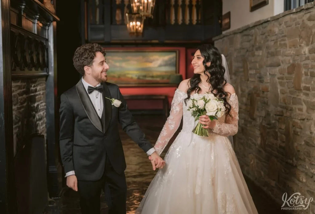 A bride and groom smiles at each other as they walk down a brick-clad corridor at Old Mill in Toronto