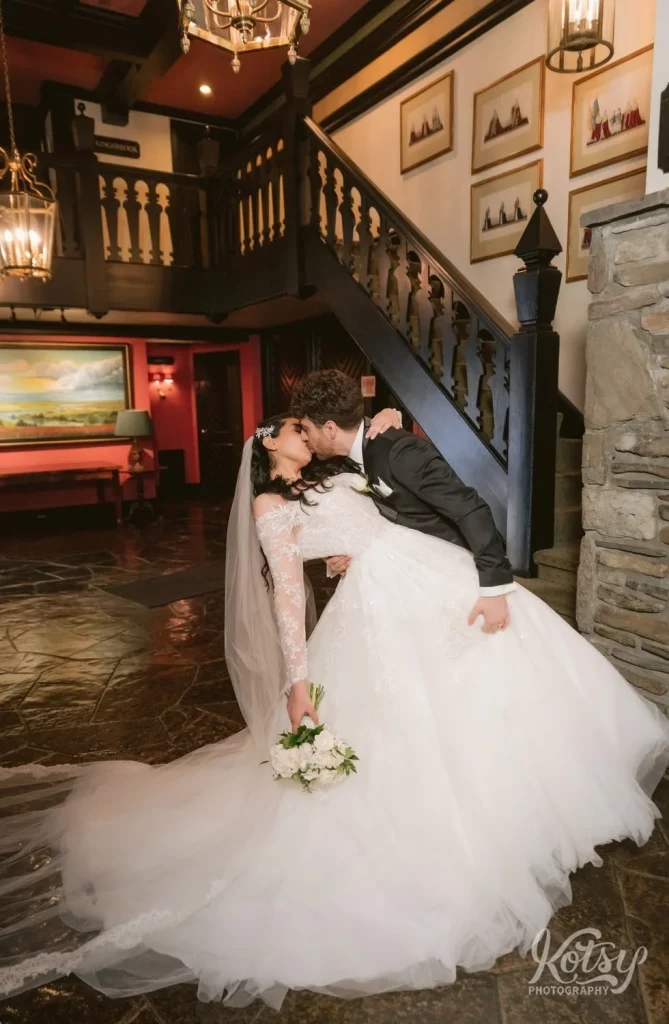 A groom dips his bride over and gives her a big kiss at the bottom of stairs inside Old Mill in Toronto