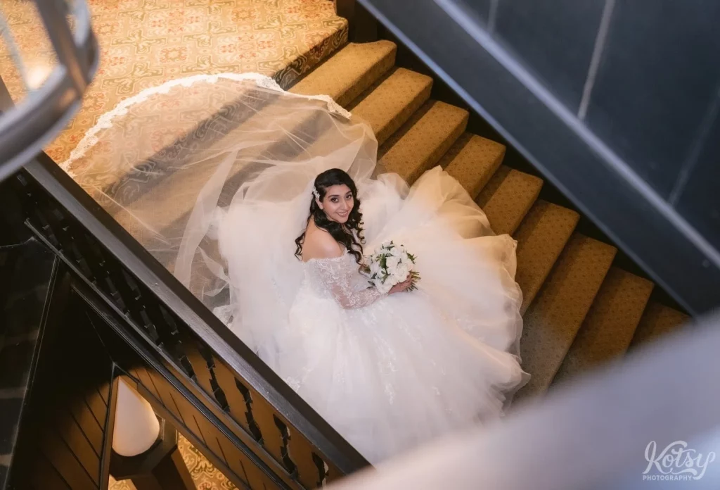 A bride looks up to the camera and smiles while sat on a large set of stairs inside Old Mill in Toronto