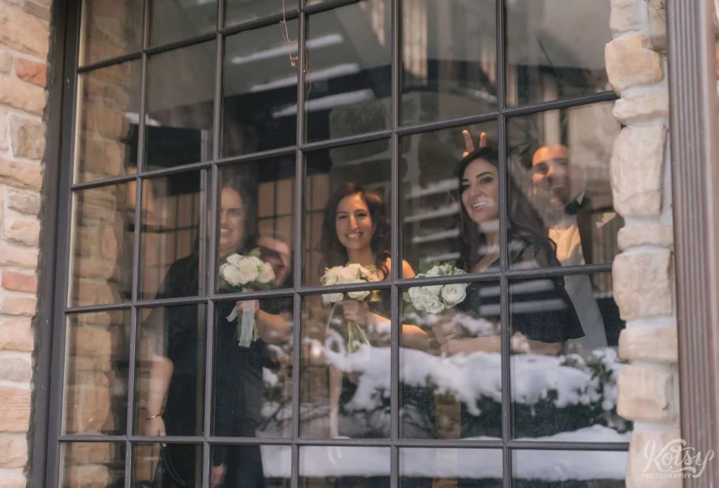 A bride and groom's wedding party smile through a window as they watch their photoshoot at Old Mill in Toronto