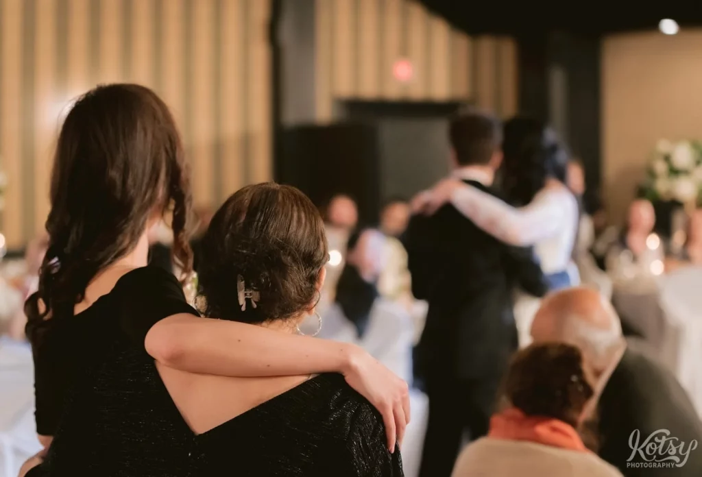 A woman with her arm around her mom watch a bride and groom enjoy their first dance at Old Mill