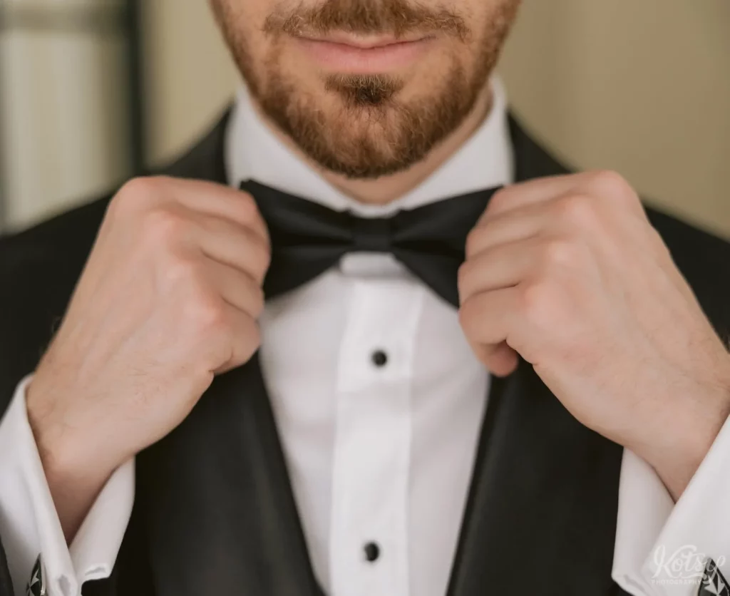 A tuxedo-clad groom pulls on both ends of his bow-tie before departing for his wedding ceremony