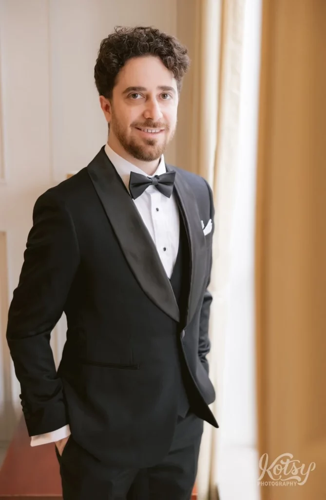 A groom in a tuxedo smiles for the camera by a window in his condo in Toronto, Canada