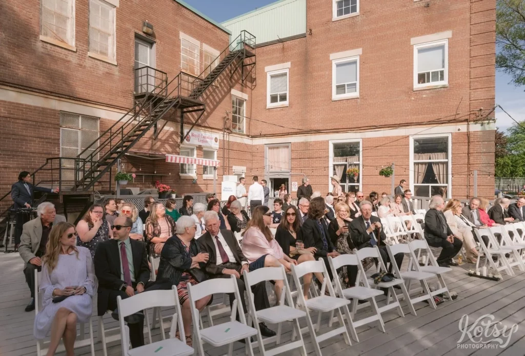 A wide shot of all the guests sitting in chairs outdoors at Royal Canadian Legion 344 awaiting the start of a wedding ceremony