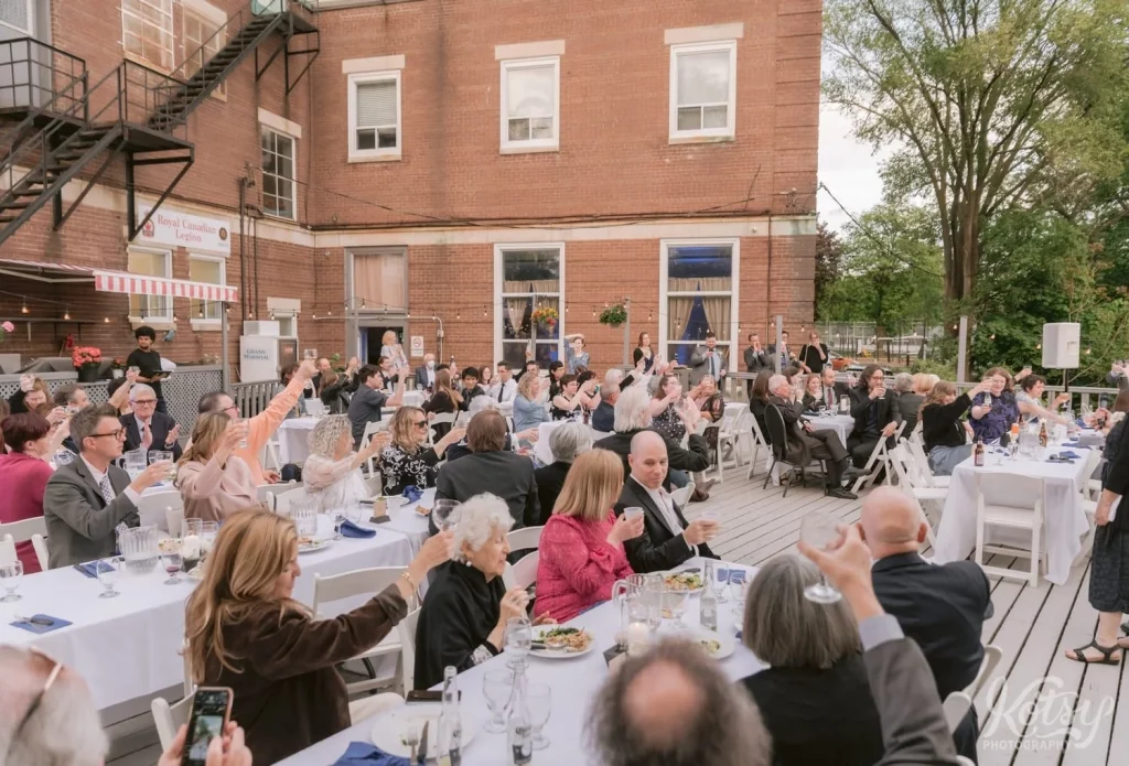 A large crowd of seated guests raise their glasses for a toast during an outdoor wedding reception at Royal Canadian Legion 344