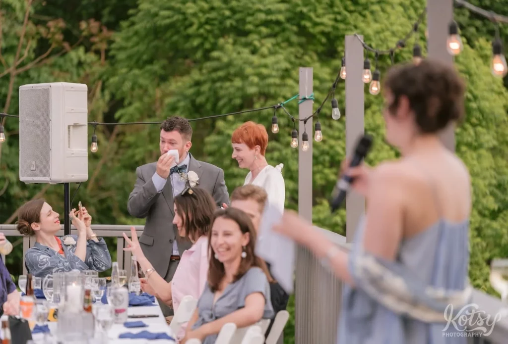 A groom wipes a tear from his eye during the maid of honour speech at his outdoor wedding reception in Toronto
