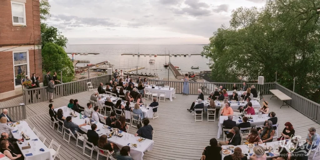 A elevated panoramic shot of a maid of honour making a speech on a large lakeside patio filled with guests at Royal Canadian Legion 344 in Toronto