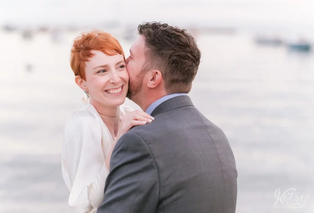 A bride smiles big as her groom whispers in her ear on a beach in Toronto